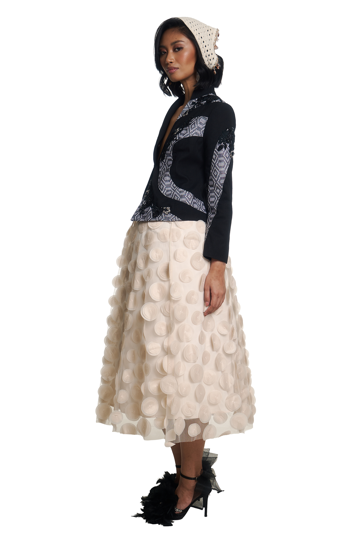 A model is shown in profile wearing a black and white patterned jacket with a cream polka-dot skirt, a cream headwrap, and black feathered high heels, set against a plain white background.