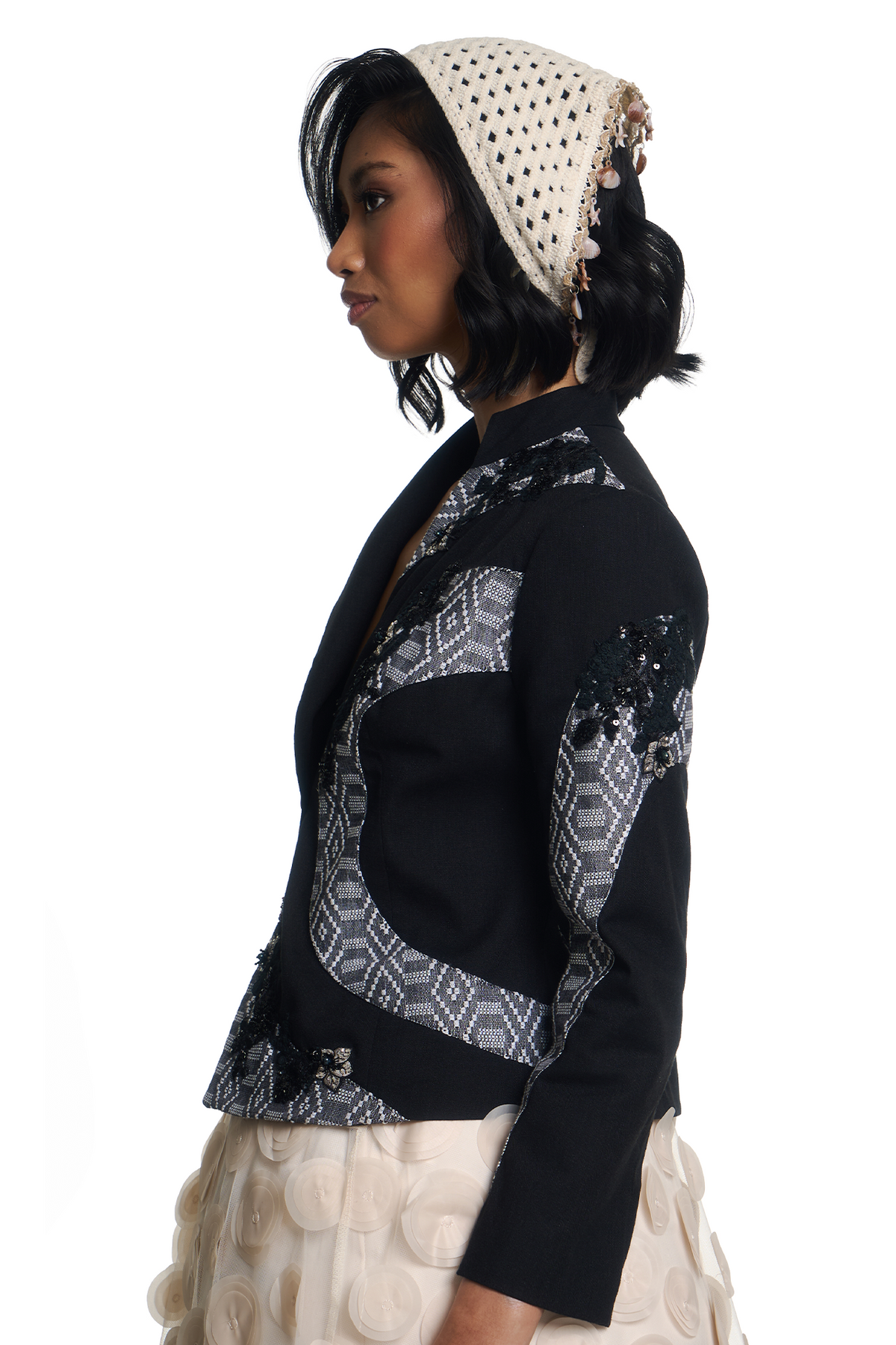A woman in profile wearing a black cropped blazer with geometric patterned panels on the front and sleeves, paired with a cream crochet head covering and a cream textured skirt, against a plain white background.