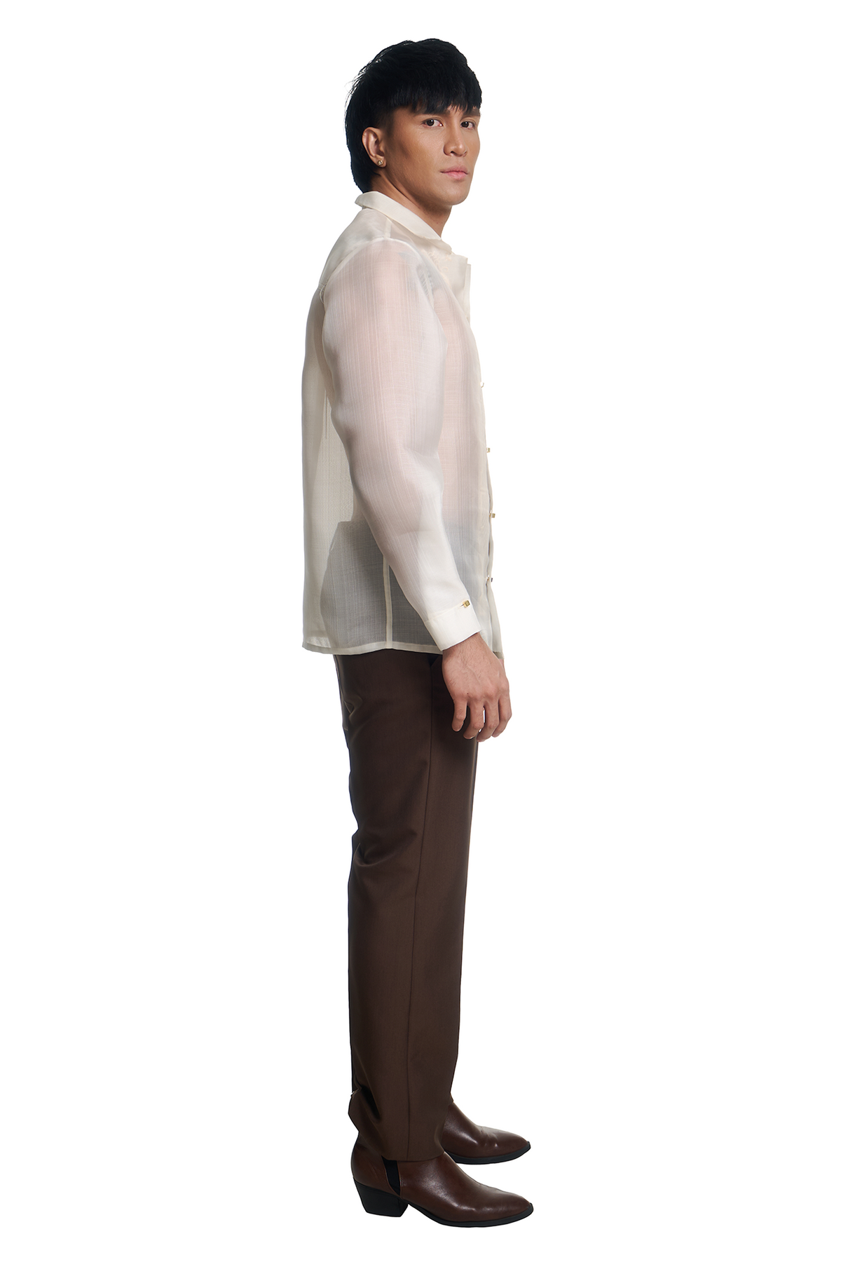 A man shown in profile wearing a white long-sleeve button-front shirt, brown trousers, and brown boots against a plain white studio background.