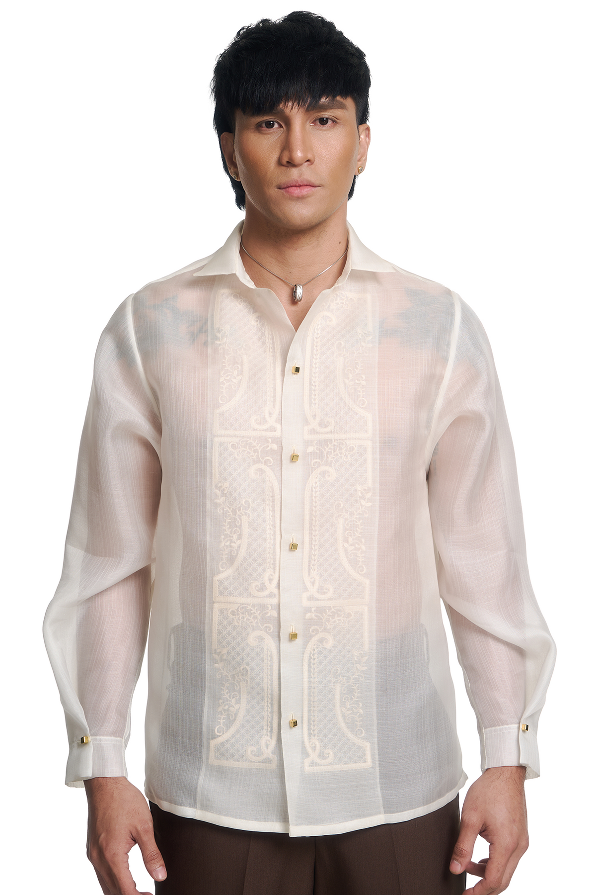 A man wearing a traditional Filipino barong shirt in off-white/cream with gold buttons and gold embroidered details on the front panel, paired with dark brown trousers, against a solid white background.