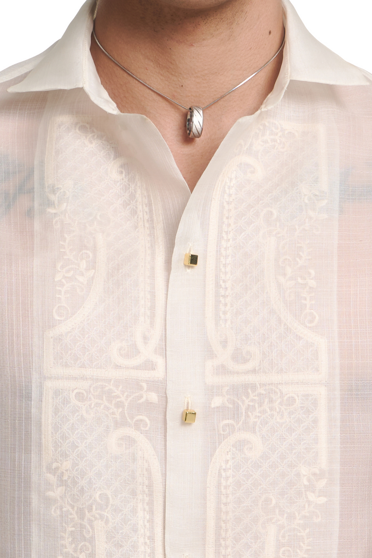 A close-up of a man wearing a white embroidered button-down shirt with gold buttons and a silver pendant necklace.