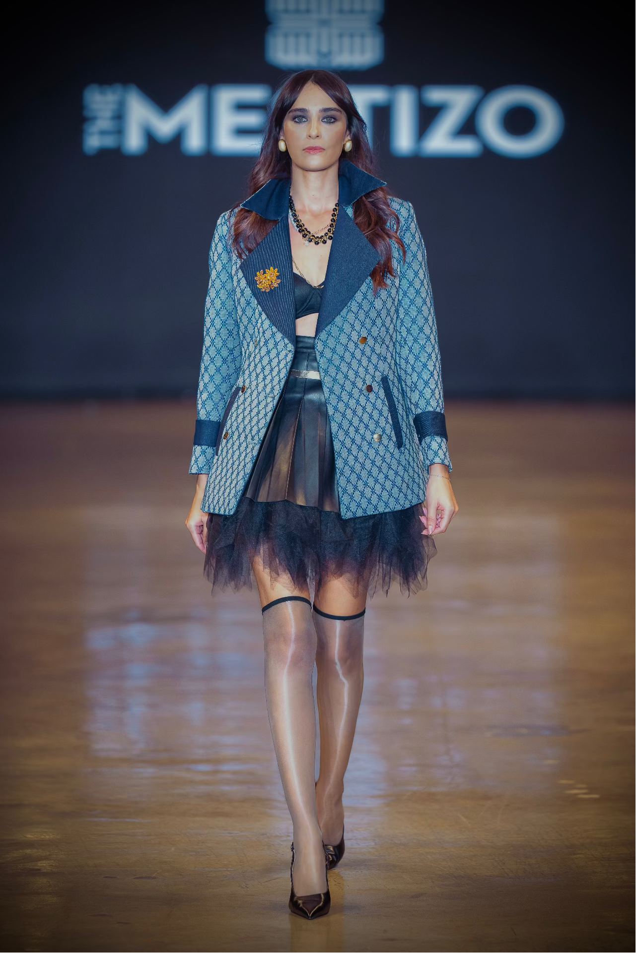 Model walking on a runway wearing a blue patterned outfit with 'The Mestizo' brand visible.