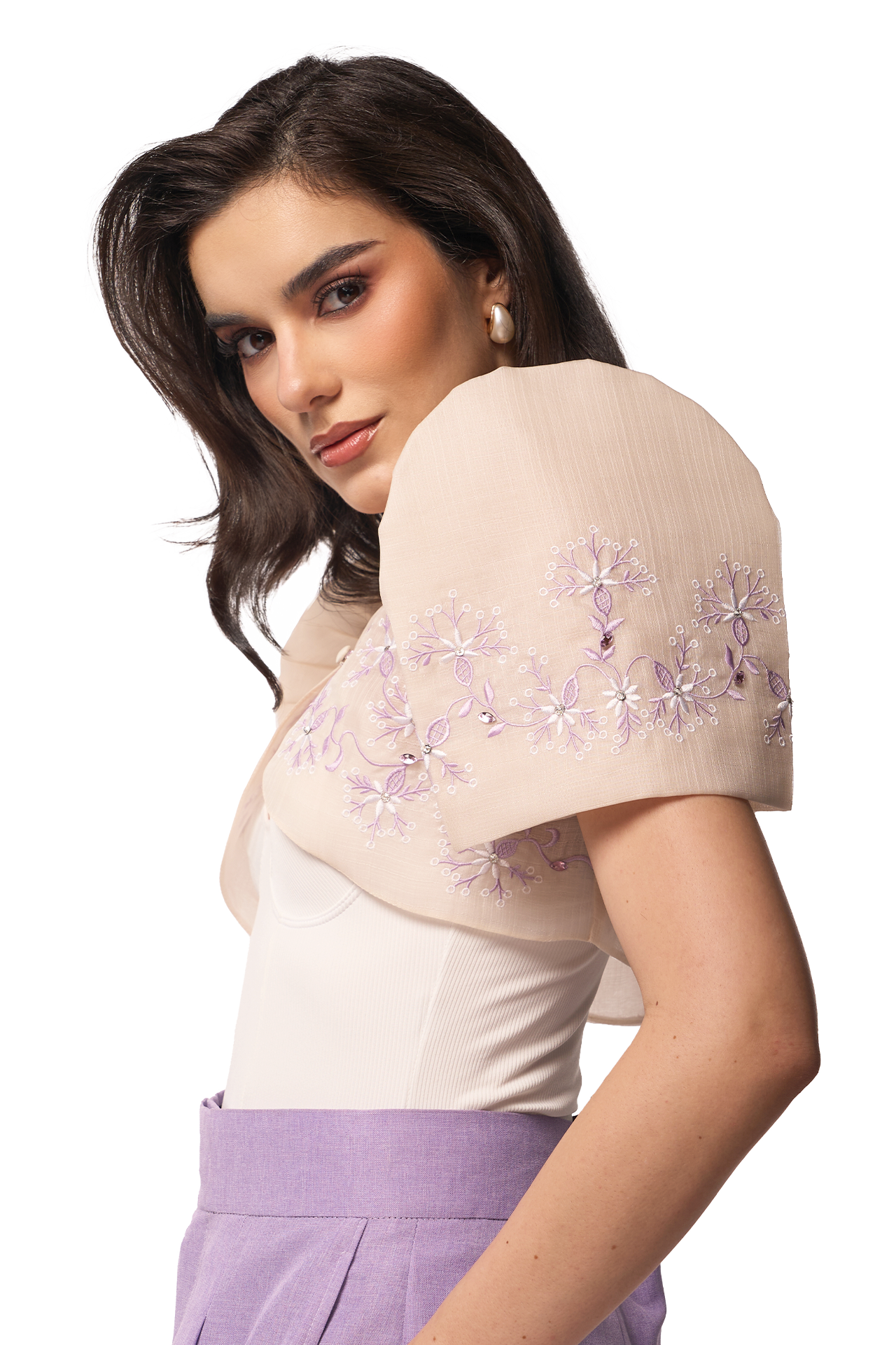 Woman wearing a beige embroidered shrug over a white blouse with purple pants on a white background