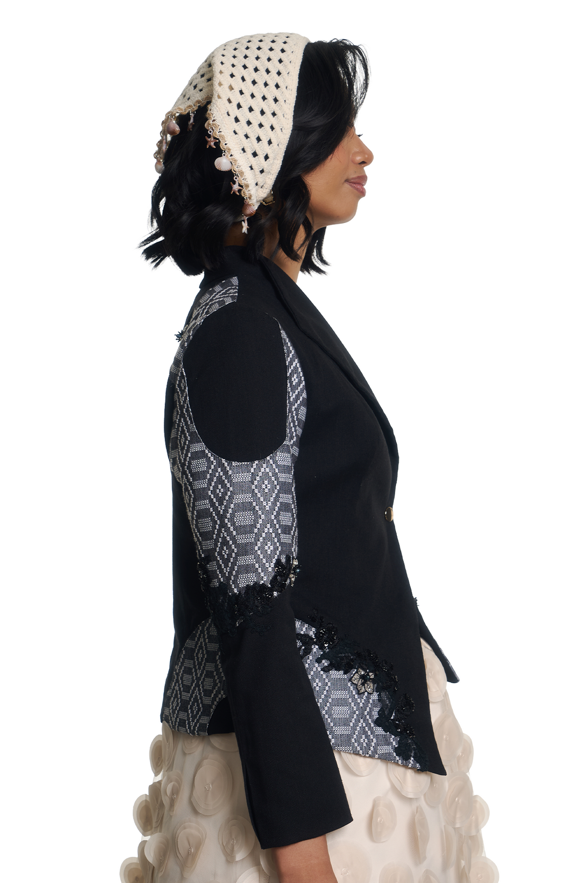 A woman shown in profile wearing a black tailored jacket with geometric patterned fabric panels on the sleeves and lower front, paired with a cream crocheted head covering and a cream textured skirt, against a plain white studio background.