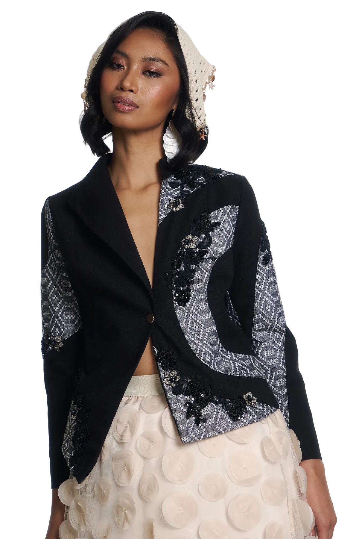 A model wearing a black cropped blazer with intricate gray and black patterned lace panels and black floral embroidery, paired with a cream 3D-embossed skirt and a matching cream headscarf.