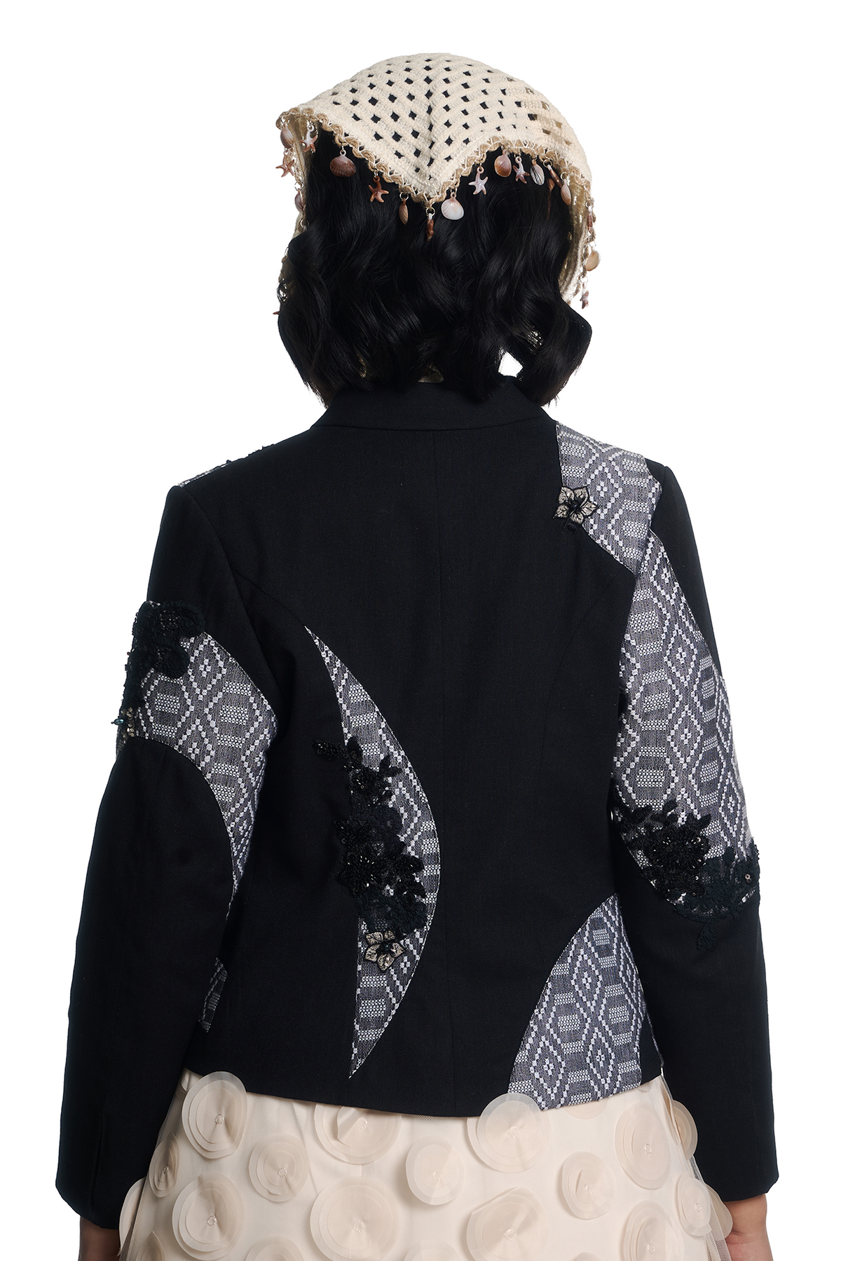 Back view of a model wearing a black cropped jacket with geometric patterned fabric panels on the sleeves and back, paired with a textured cream skirt and a matching cream beanie.