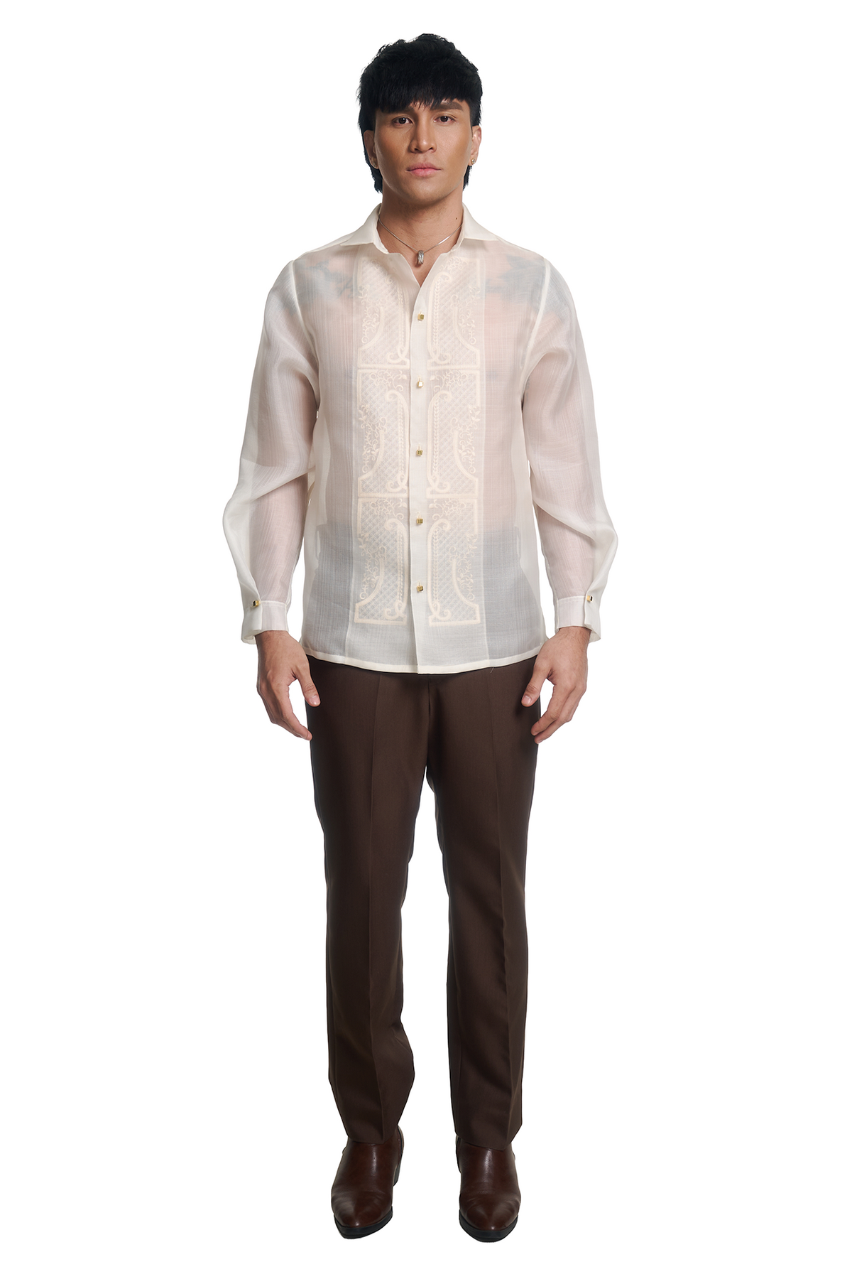 A man modeling a traditional Filipino barong shirt in off-white with cream embroidery, paired with dark brown trousers and brown leather shoes, against a solid white background.