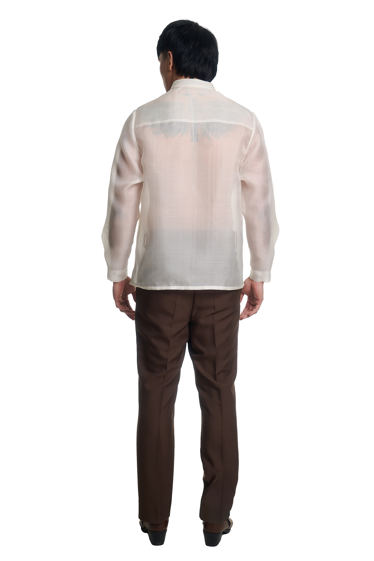 A man viewed from the back wearing a white semi-sheer Philippine barong shirt with a subtle gray wing motif on the upper back, paired with dark brown trousers and black shoes, against a solid white background.