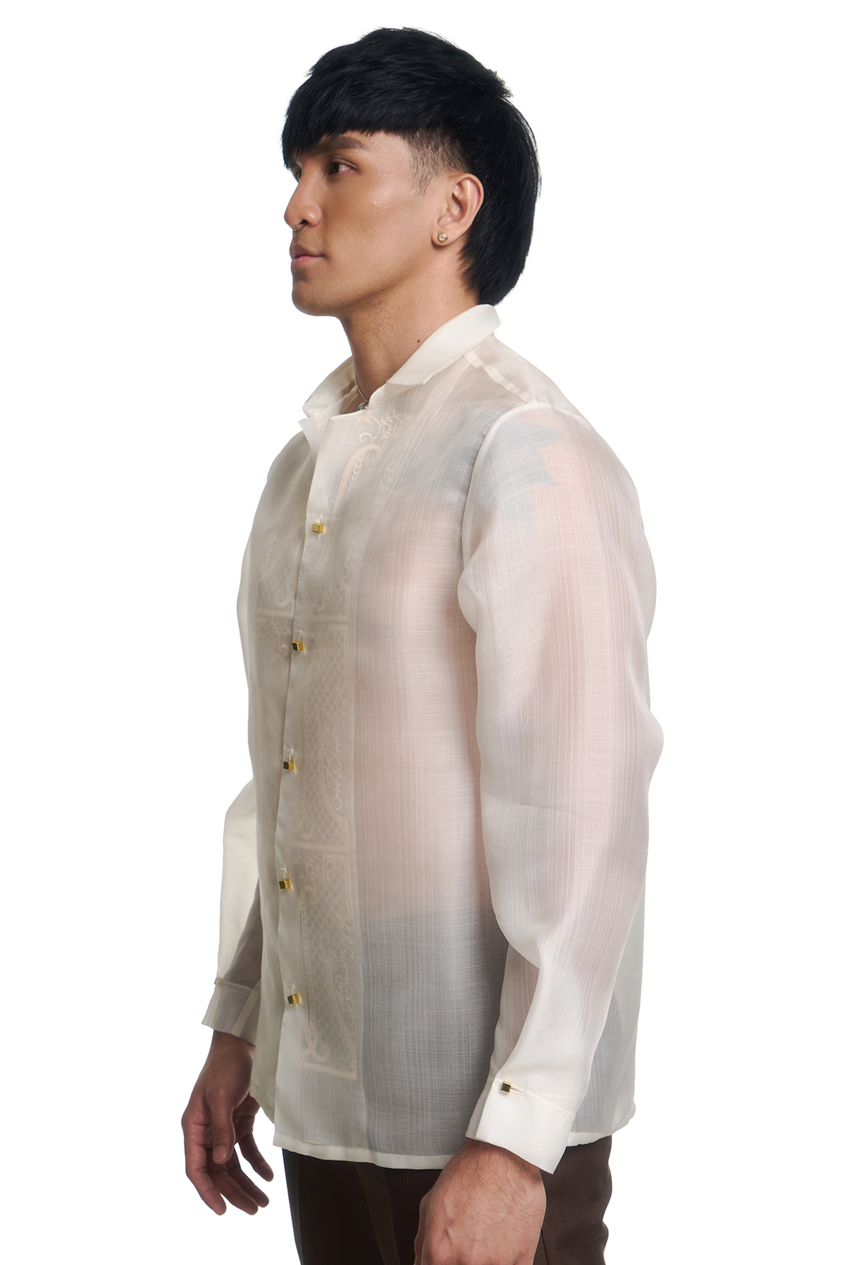 A man shown in profile wearing a lightweight, semi-sheer barong shirt in a warm cream or off-white color with gold buttons. The background is a clean, solid white.