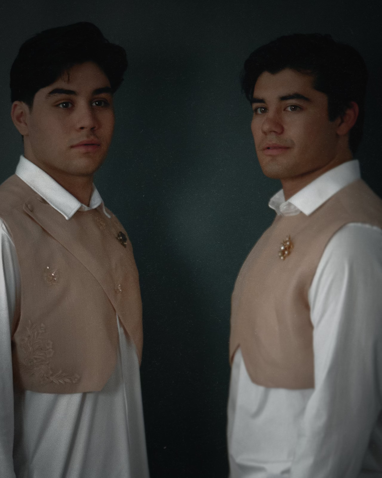 Two men wearing matching beige embroidered waistcoats over white collared shirts, posed against a dark studio background.